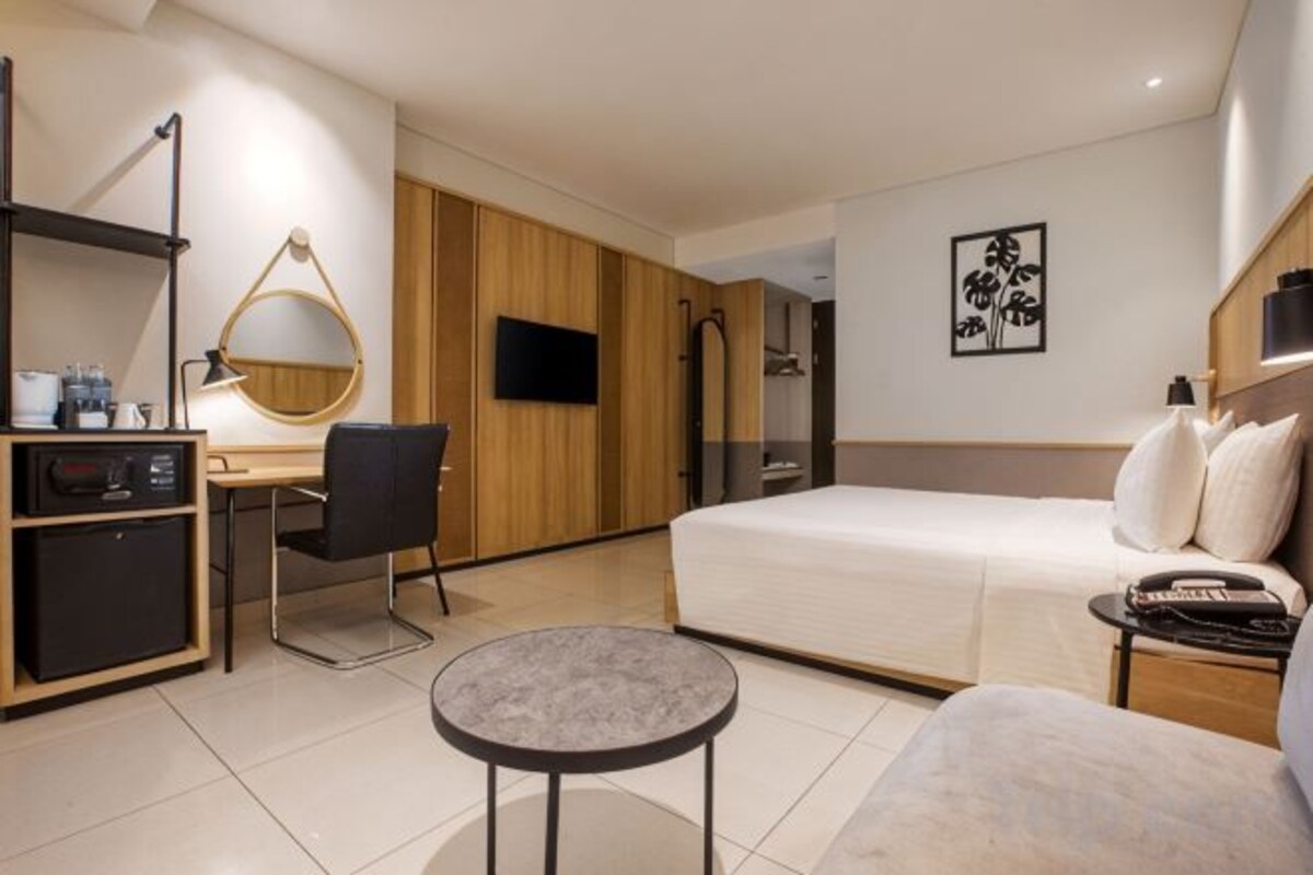 Habitación de hotel con una cama, un escritorio con silla, una mesita redonda, un televisor y un armario.