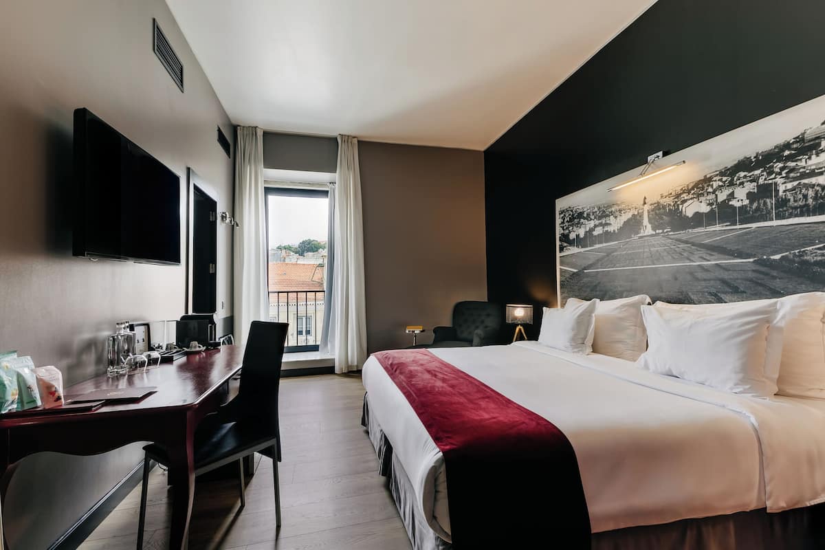 Ein Hotelzimmer mit einem großen Bett, einem Schreibtisch mit Stuhl, einem an der Wand montierten Fernseher und einem großen Fenster mit Blick auf Gebäude.