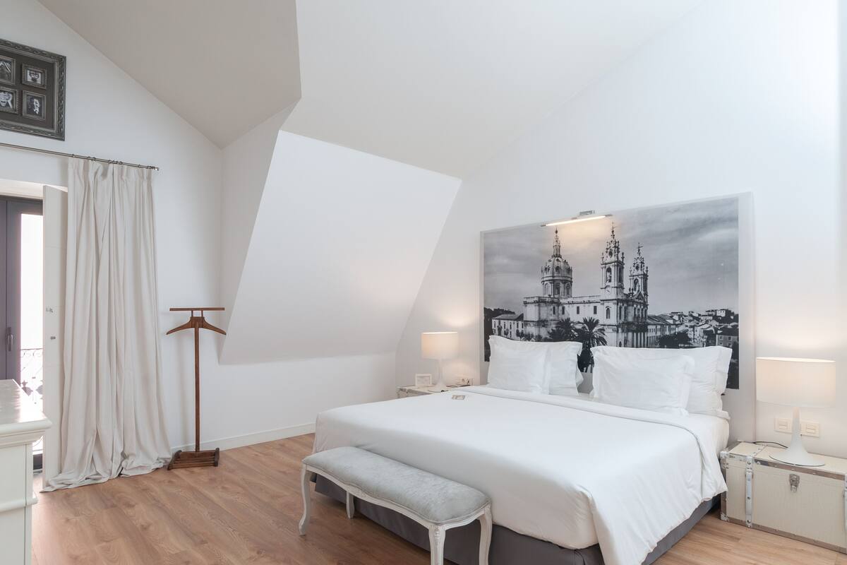 Ein Schlafzimmer mit einem Bett, einer Bank, Nachttischen, einem Kleiderhaken und einem großen schwarz-weißen Stadtbild an der Wand.