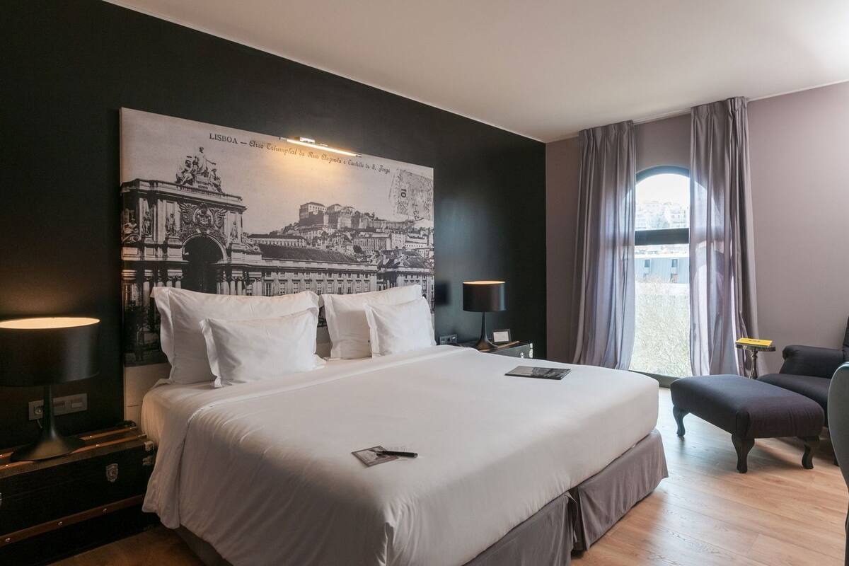 Ein Hotelzimmer mit einem großen Bett, einem Sessel, einem Schreibtisch, einer Lampe und Blick auf die Stadt.
