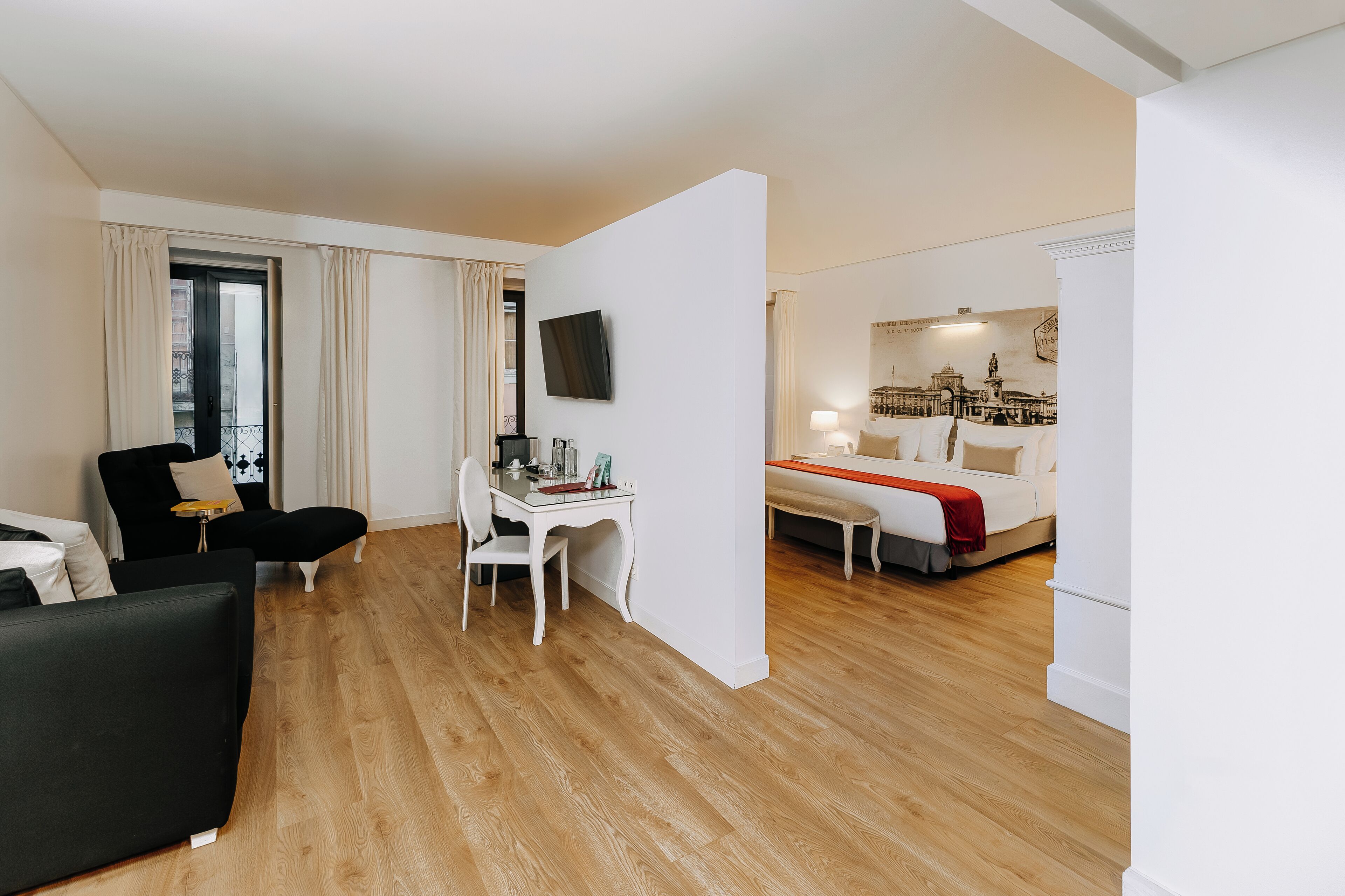 Moderni hotellihuone, jossa on puulattia, musta sohva, valkoinen työpöytä ja punaisella peitolla varustettu sänky.