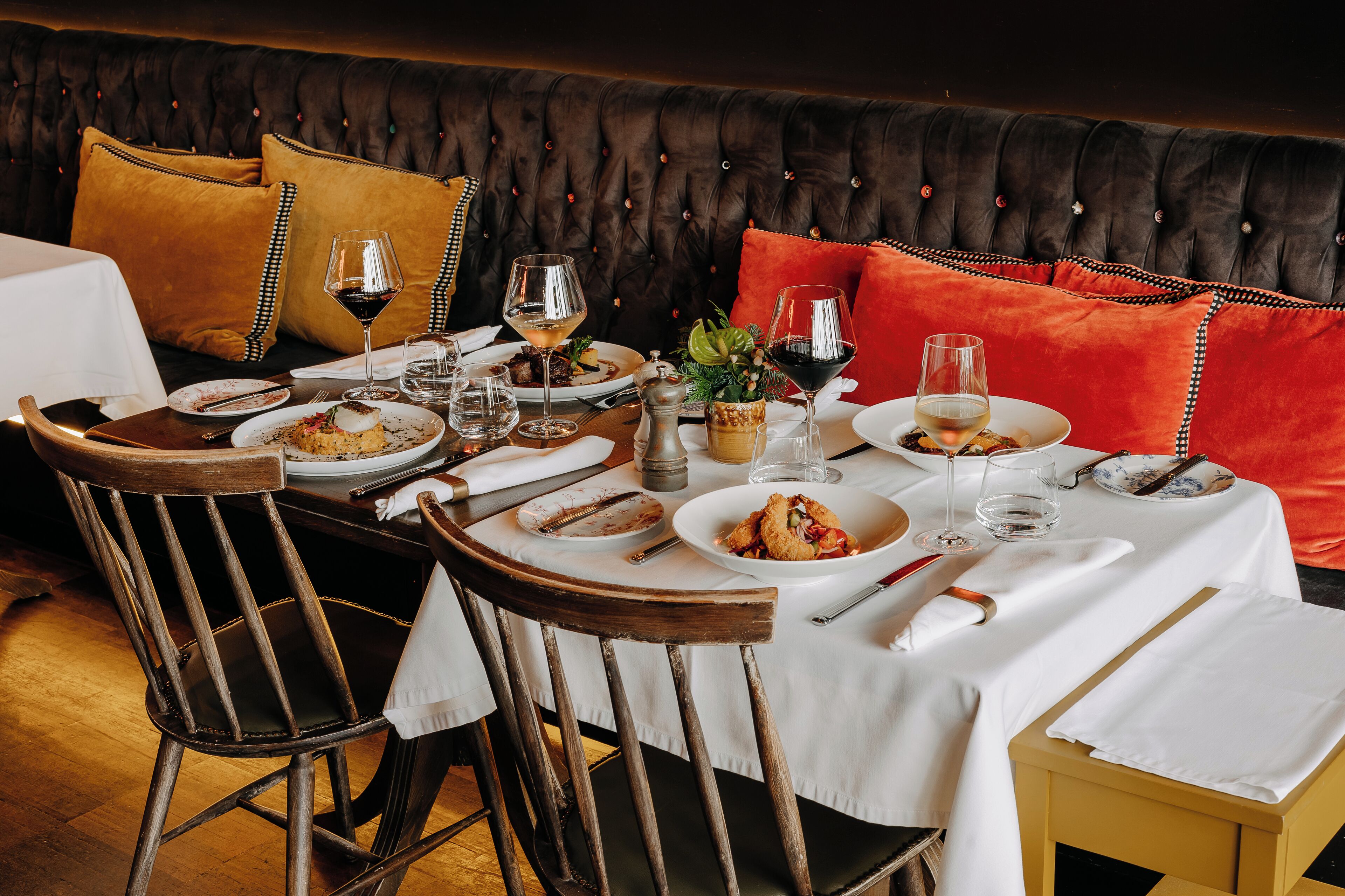 Une table à manger dressée avec des assiettes, des verres et des couverts, un banc rembourré avec des coussins et une petite plante.