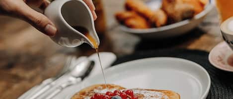 Desayuno con tortitas cubiertas de frutos rojos y jarabe, un bol de croissants y un vaso de zumo de naranja sobre una mesa de madera.