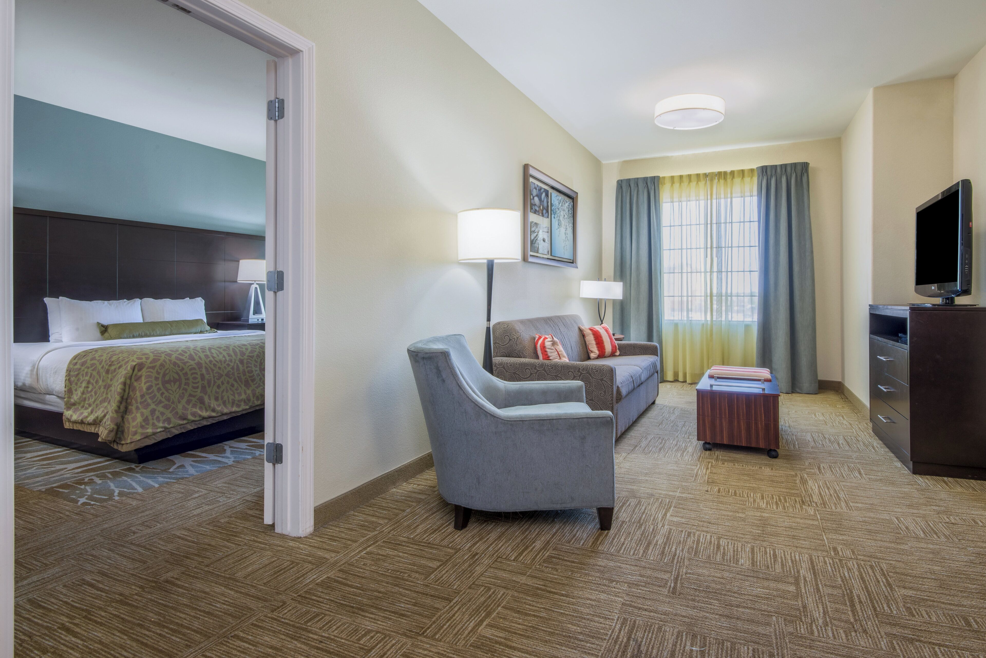 Habitación de hotel con cama, sofá, escritorio, televisor y ventana con cortinas.