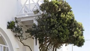 A narrow, white-walled corridor with a tiled floor, leading to a covered area with a white railing and a tree.