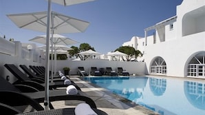 A poolside area with lounge chairs and umbrellas, a clear blue pool, and white buildings with arched windows.