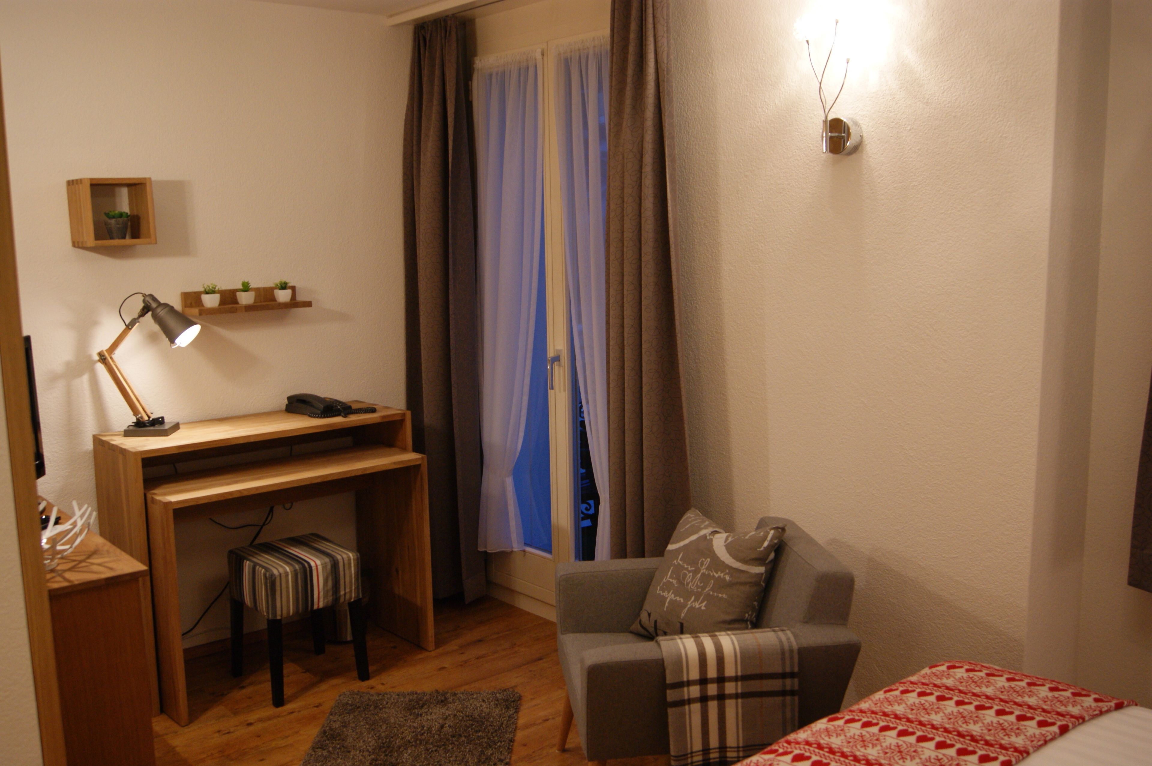 Una habitación compacta con escritorio, una silla, una cama y un taburete pequeño.