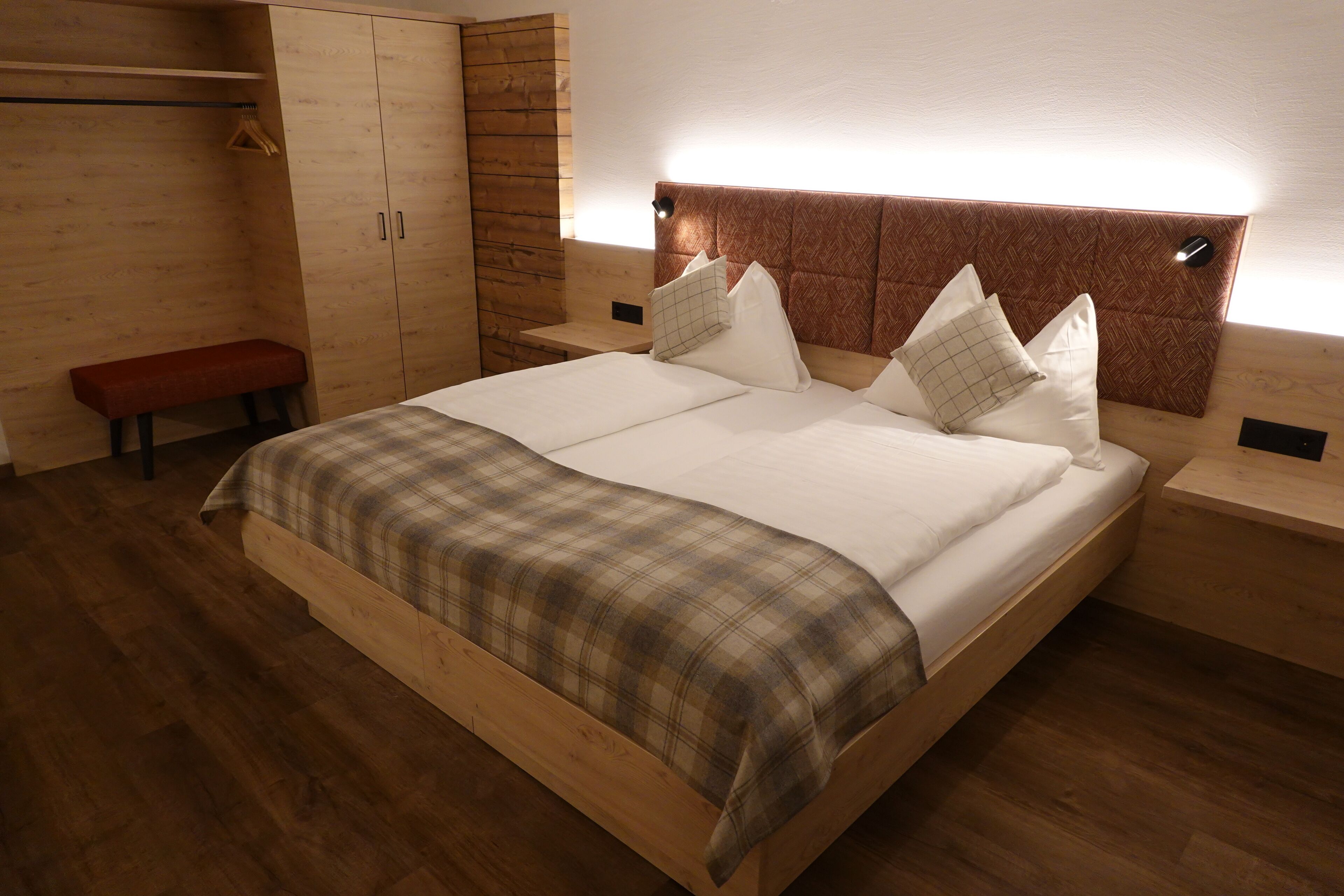 Ein hölzernes Bett mit karierter Decke, ein Kopfteil mit Lampe sowie ein Holzschrank mit Sitzbank.