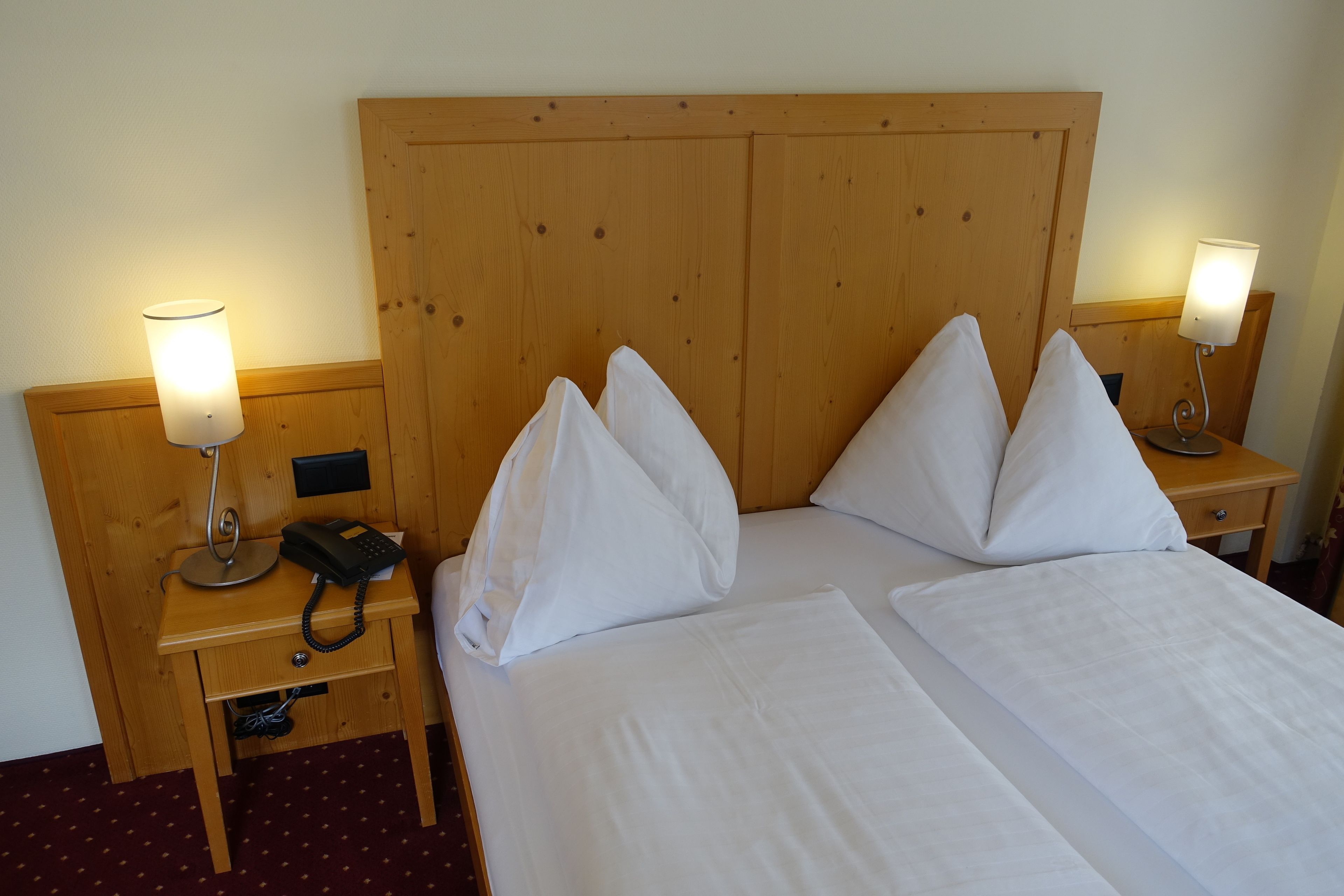 A hotel room with a wooden headboard, two bedside tables with lamps, a telephone, and neatly made beds with white linens.