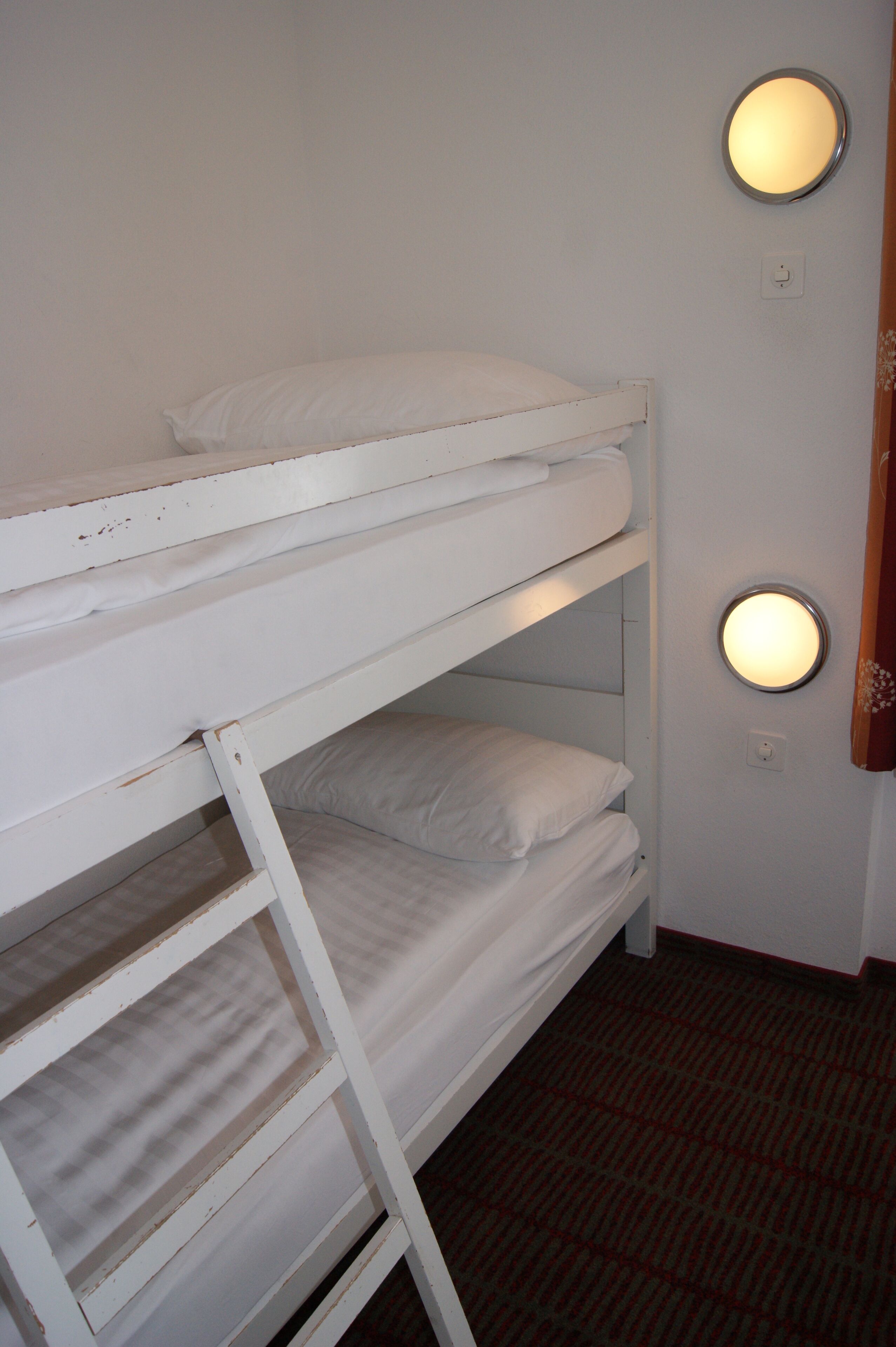 Een stapelbed met twee verhogingen, elk voorzien van kussens en dekens. Aan weerszijden van het bed zijn er twee wandlampen.