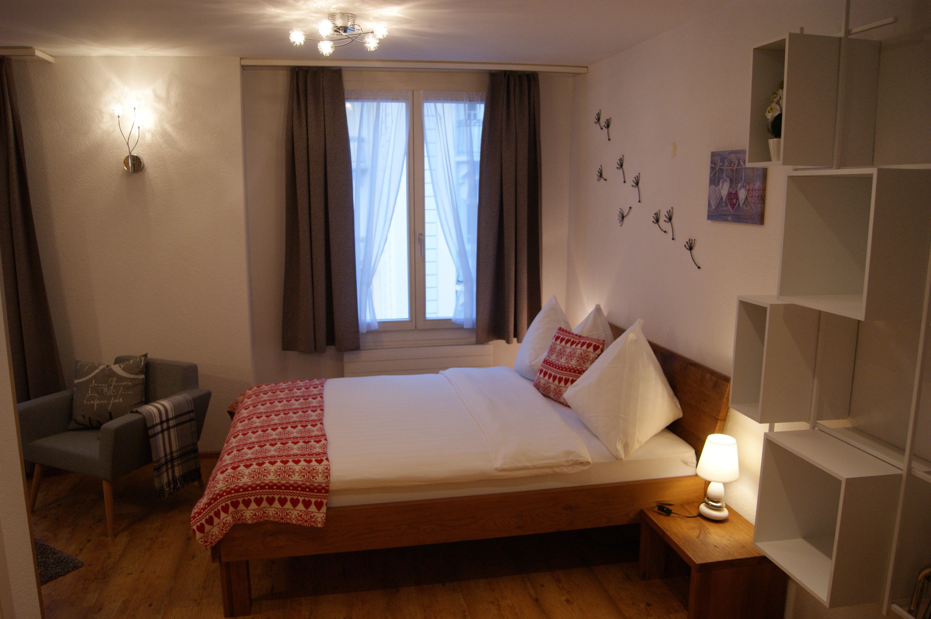 Un dormitorio con una cama de madera, una mesita de noche con lámpara, una silla con manta y una ventana con cortinas.