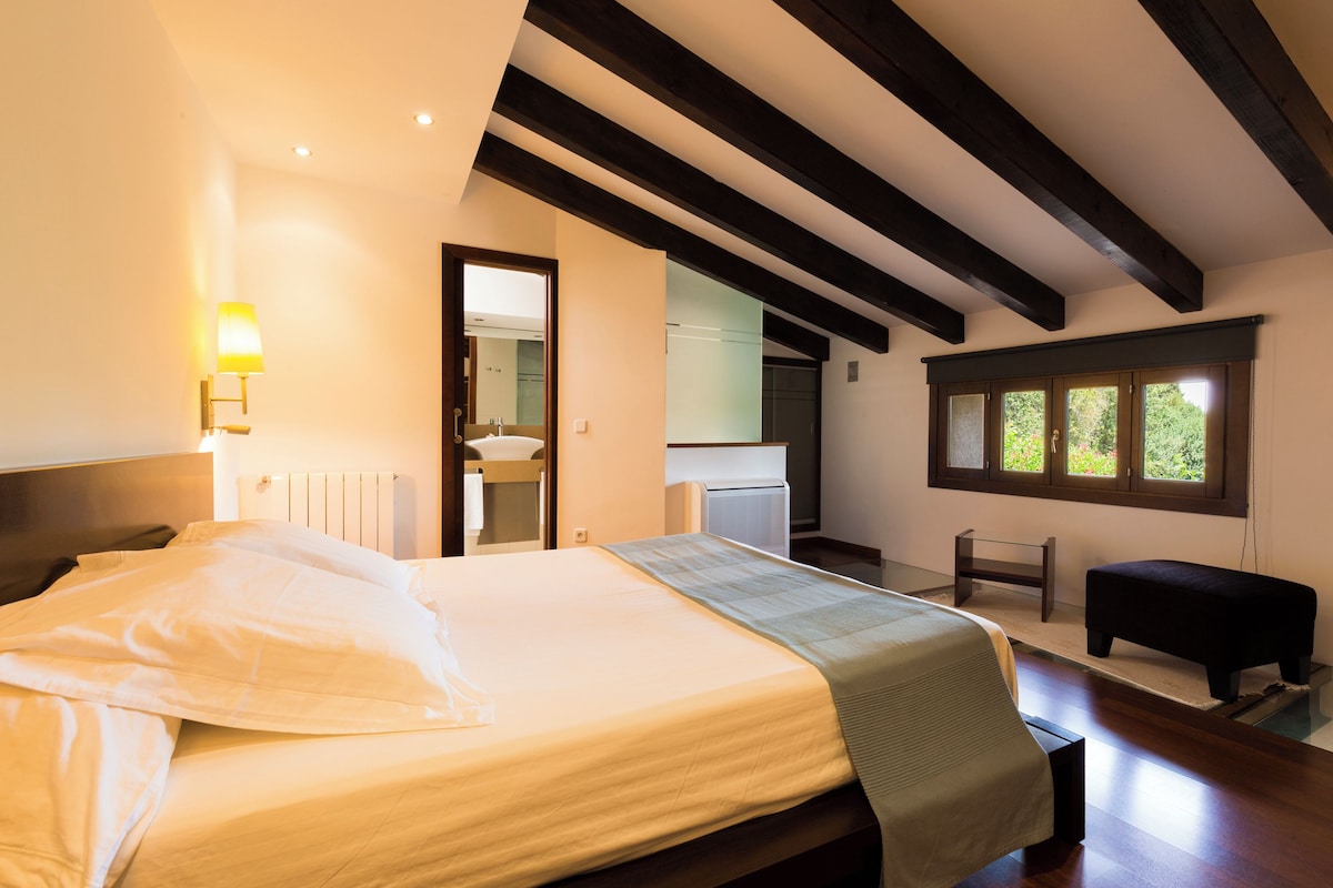 A bedroom with a large bed, a bedside table, a bench, a window, and a view of the outdoors.