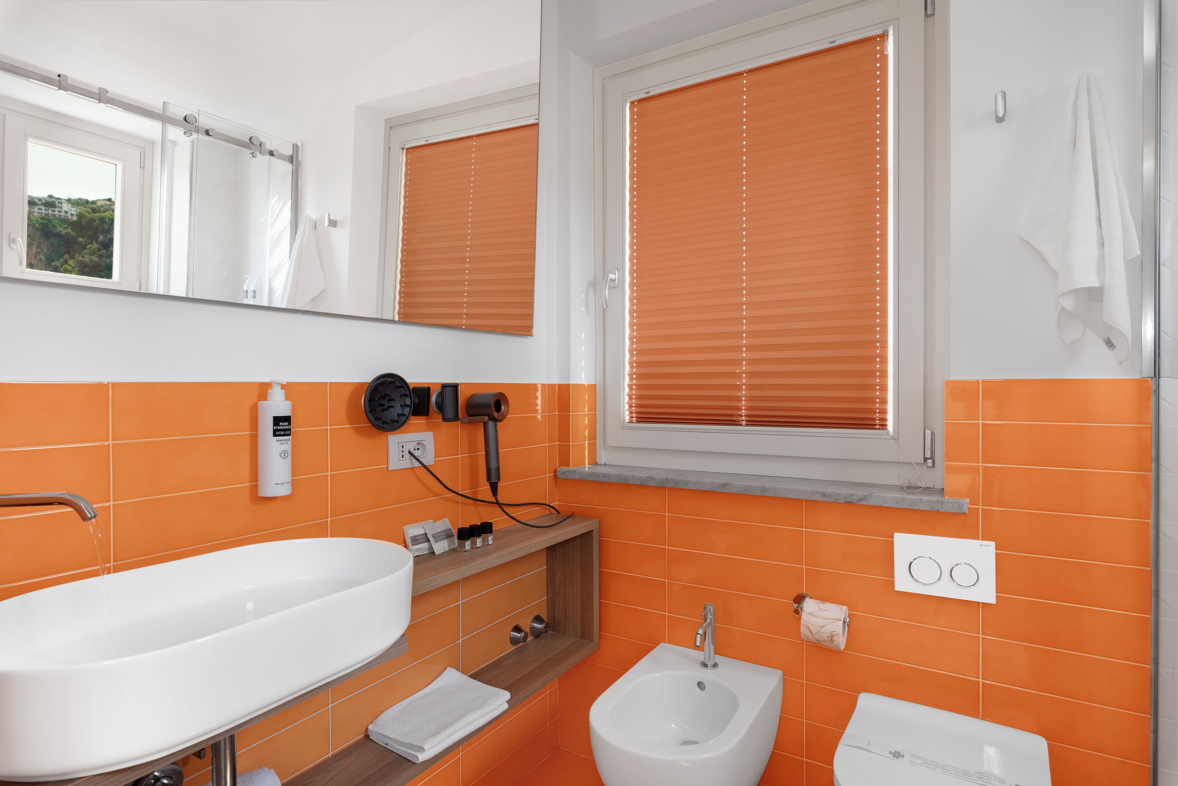 Une salle de bain avec un lavabo blanc, un bidet et des toilettes, des carreaux orange et une fenêtre avec des stores.