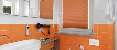 Une salle de bain avec un lavabo blanc, un bidet et des toilettes, des carreaux orange et une fenêtre avec des stores.