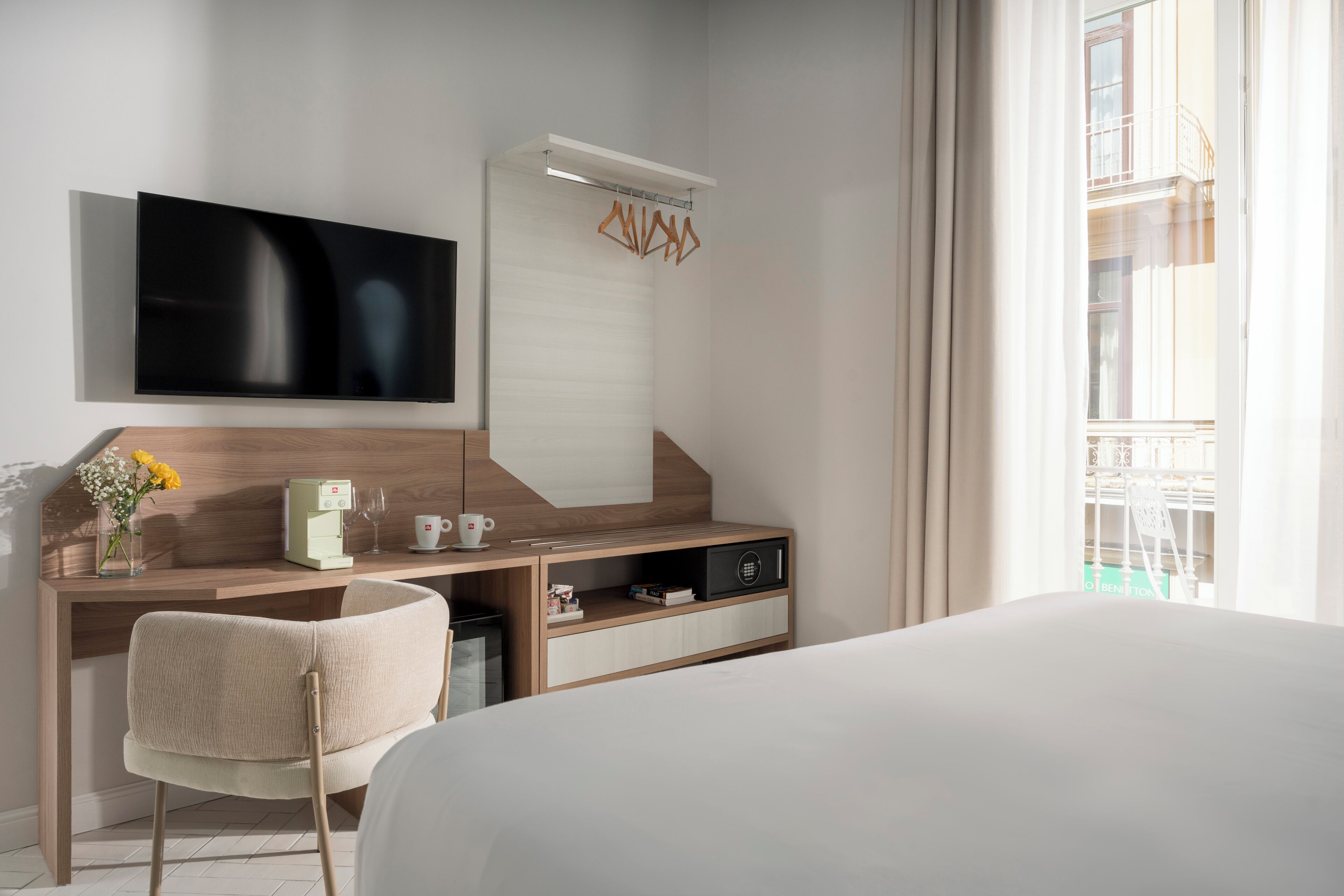 Habitación de hotel con cama, escritorio con televisor de pantalla plana, silla, cafetera y ventana con vista a un balcón.