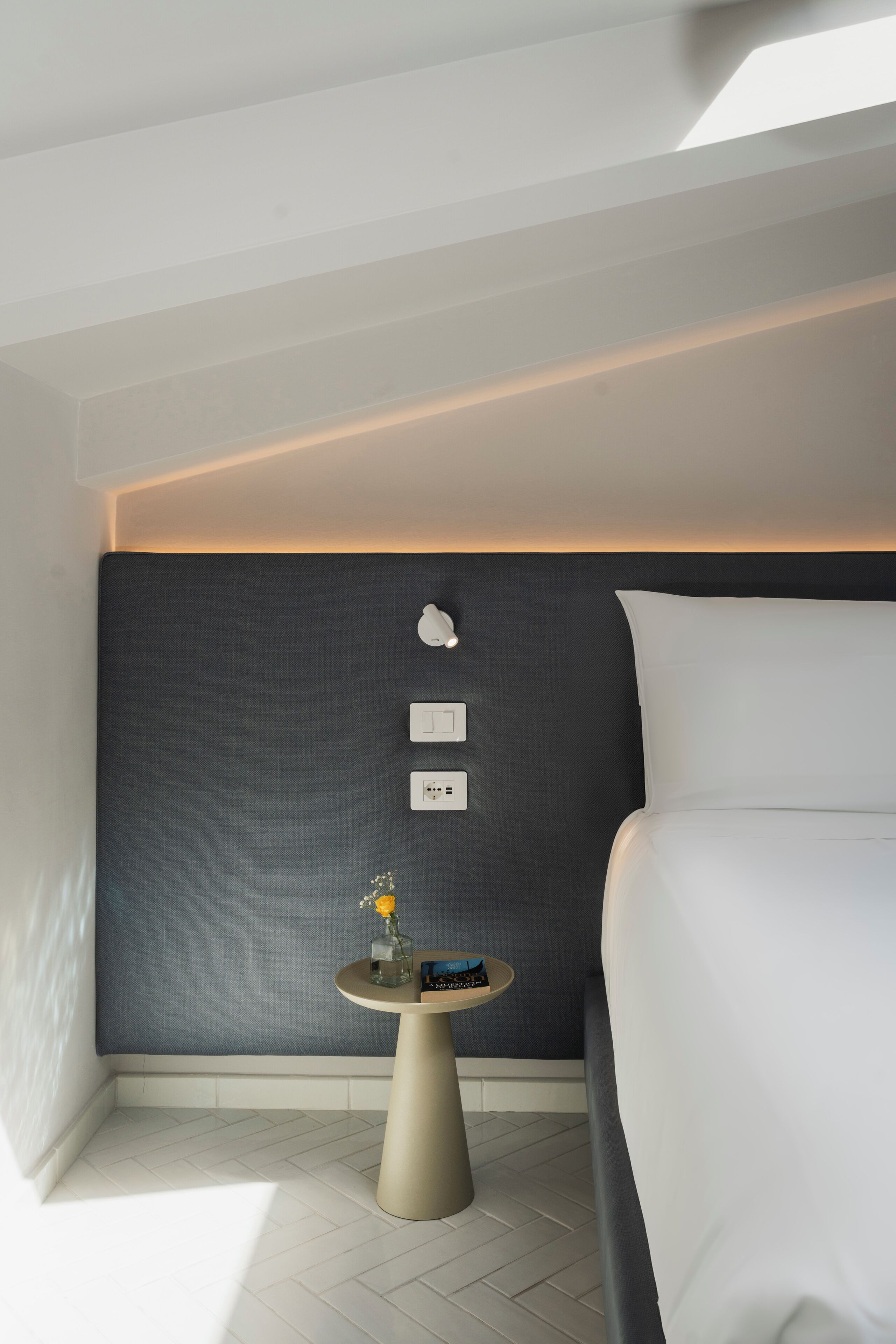 A modern hotel room with a bed, a small round table with a vase, and wall-mounted light switches.