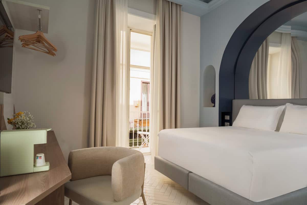 Habitación de hotel con una cama, un escritorio con silla, una lámpara colgante y vistas al exterior.