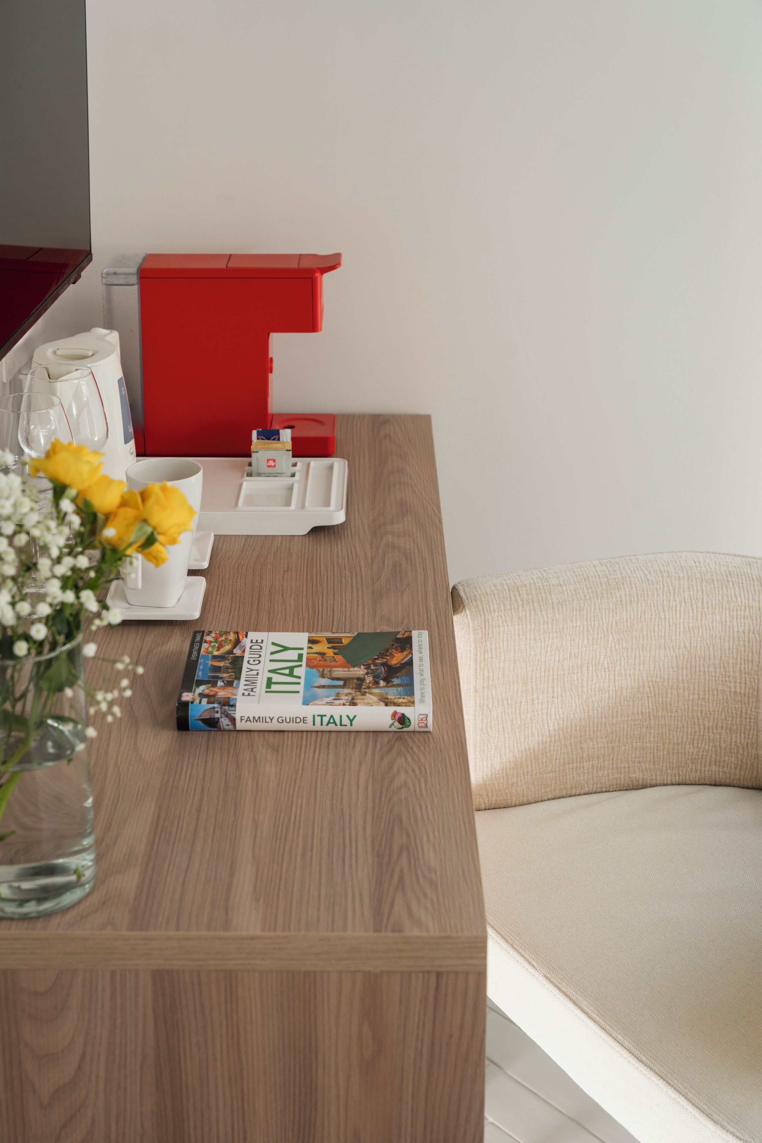 Ein Holztisch mit einer Zeitschrift, einer Vase mit Blumen und einem roten Gegenstand an einer weißen Wand.