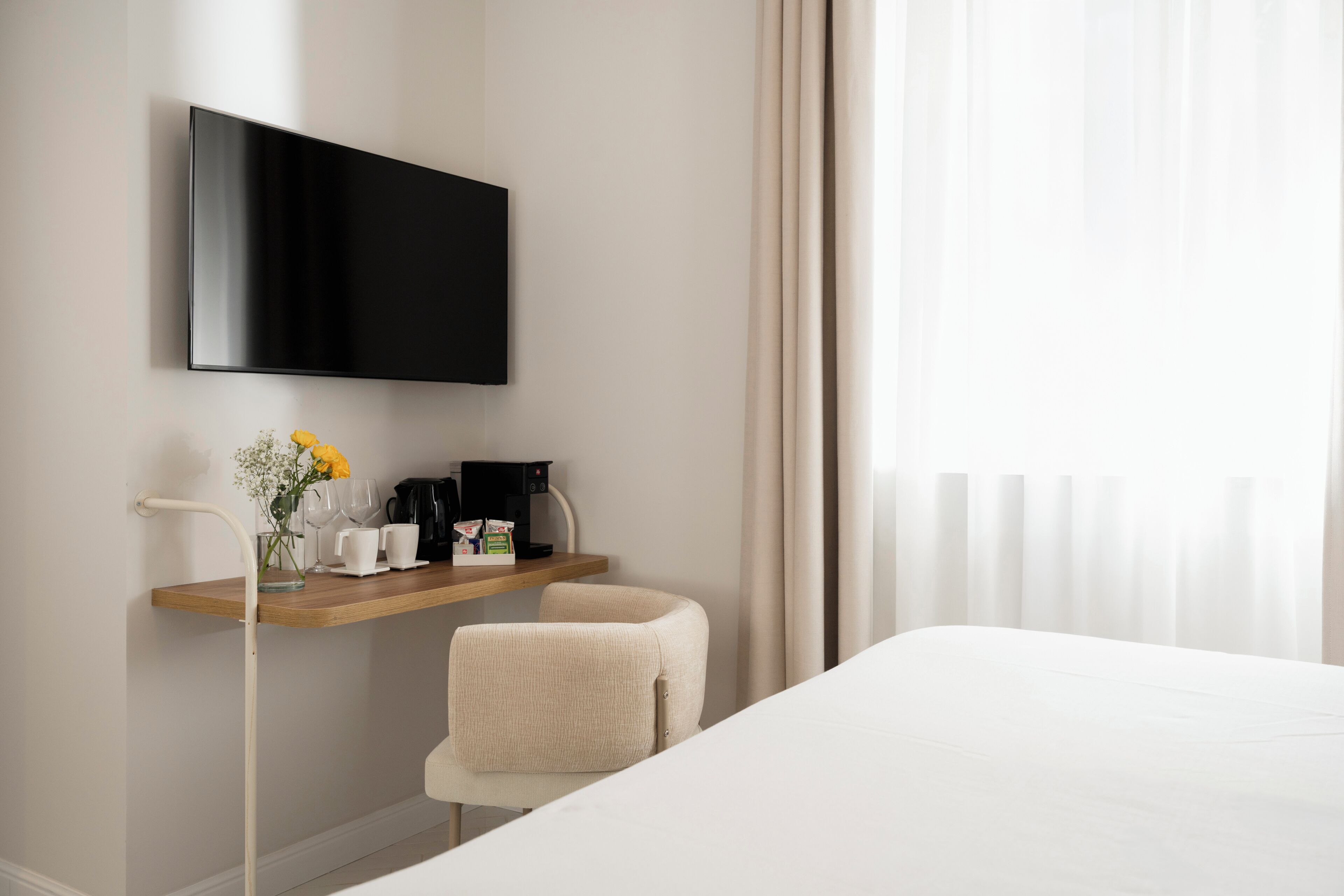 Et hotelværelse med en seng, et fjernsyn ophængt på væggen, et lille bord med kaffemaskine, kopper og en vase med blomster.