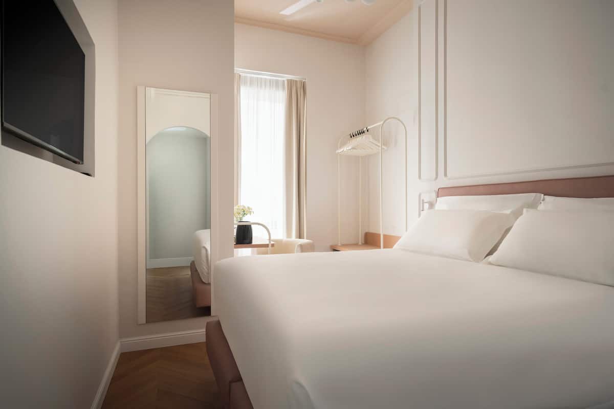 Habitación de hotel con una cama grande, una mesita de noche, un espejo, una lámpara colgante y una ventana con cortinas.
