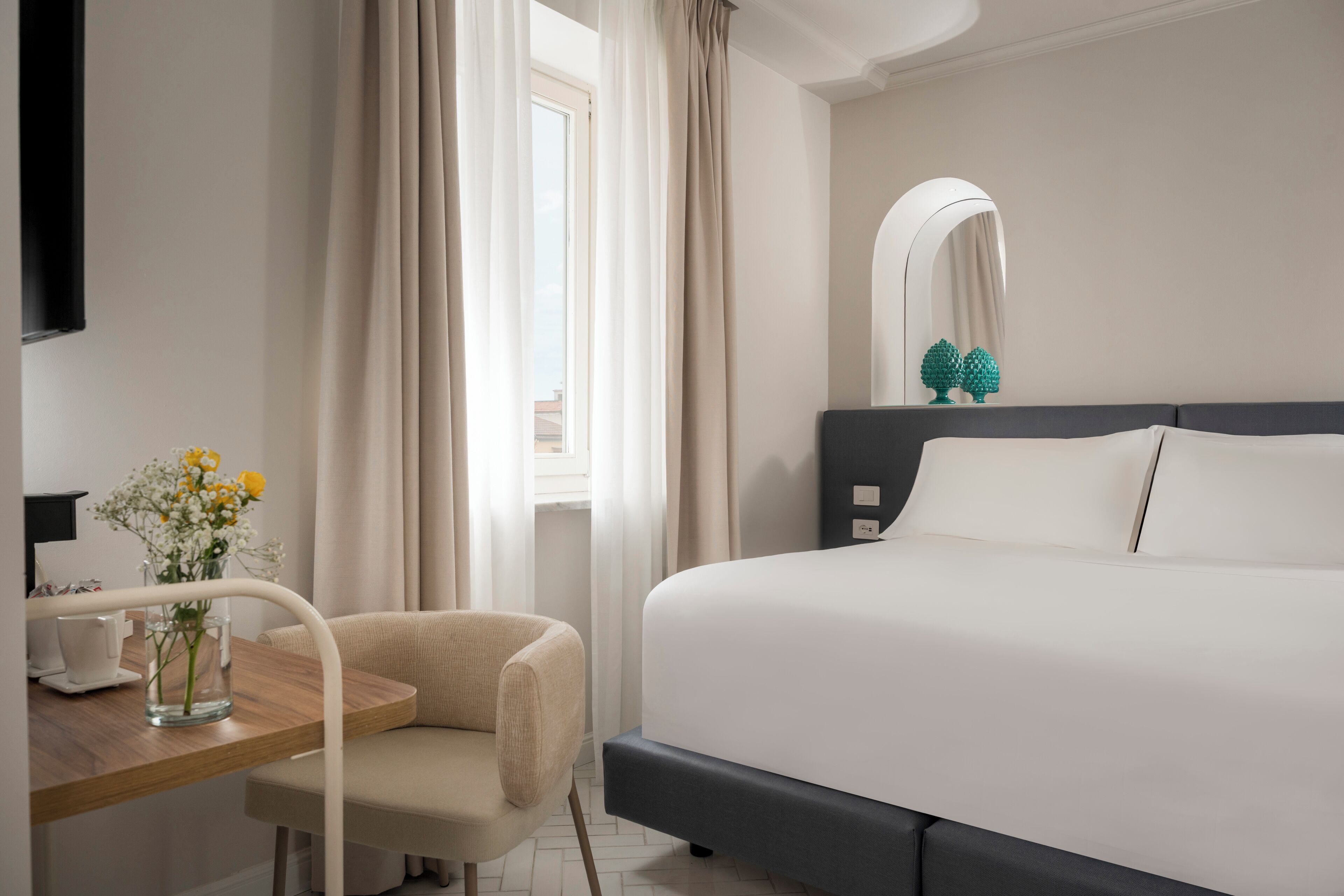 Une chambre d’hôtel avec un lit, une chaise, un bureau agrémenté d’un vase de fleurs, un miroir et une fenêtre avec des rideaux.