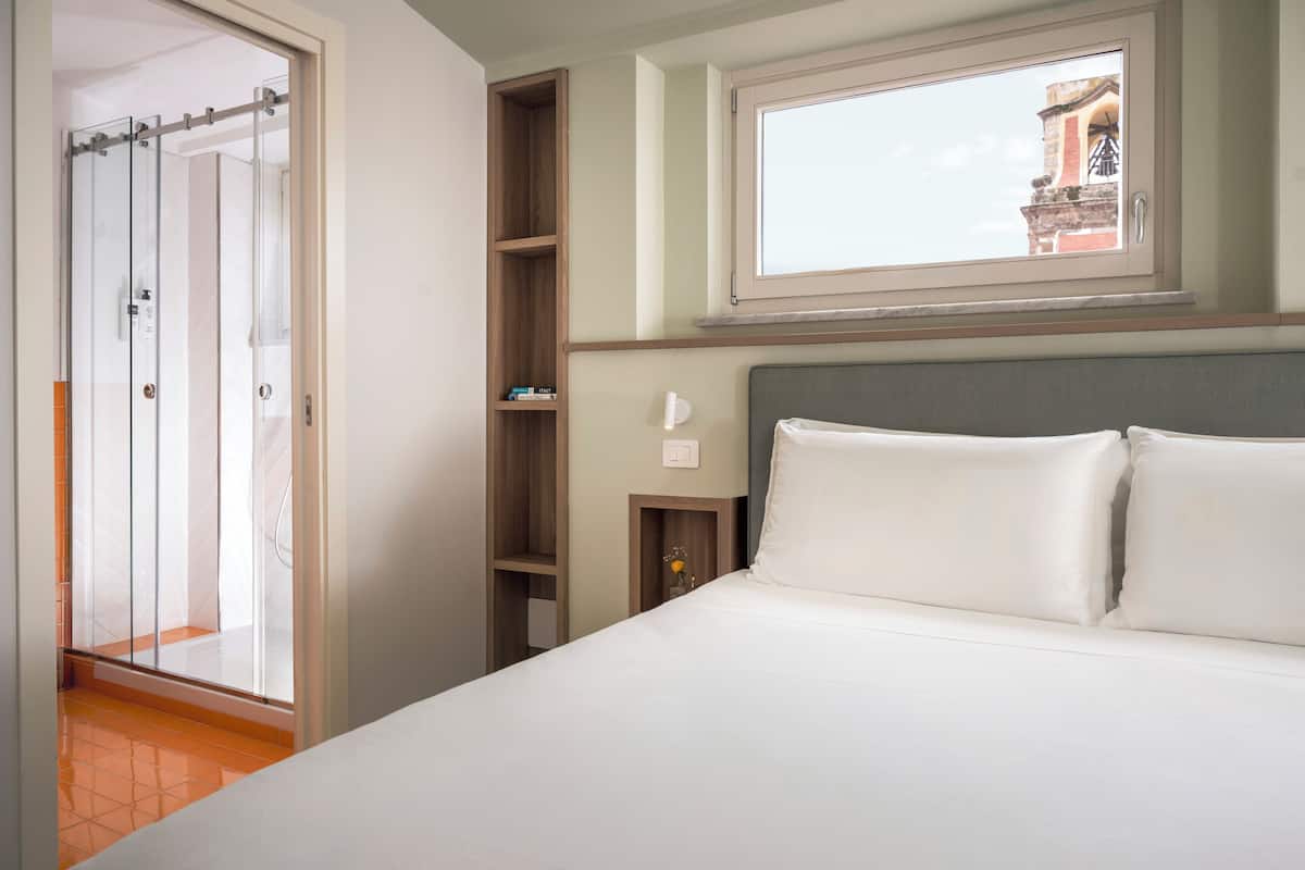 Un cuarto de hotel con una cama, una ventana con vistas, un estante de madera y una puerta que da a un baño.