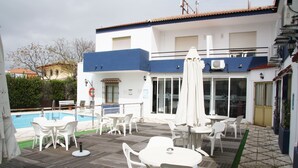 A poolside area with white plastic chairs and tables, a covered seating area, and a building with a blue and white facade. - Hotel Matalascañas (Almonte)