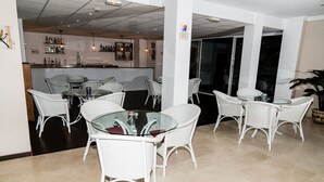 A bar area with white wicker chairs and a glass table, a tiled floor, and a bar stocked with bottles. - Hotel Matalascañas (Almonte)