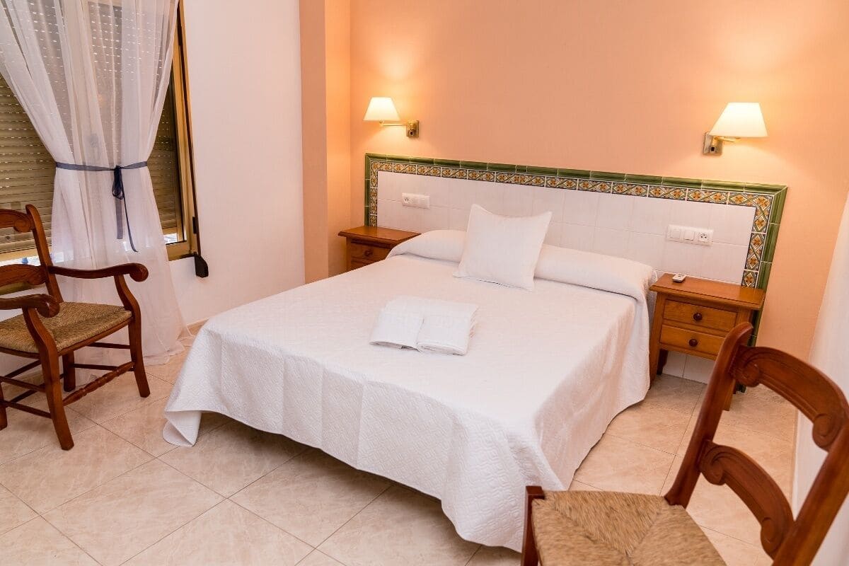 Una cama bien hecha con ropa blanca, dos mesitas de noche con lámparas y dos sillas de madera.