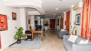 A modern living room with a grey sofa, a wooden coffee table, and a television. There is a potted plant and a red curtain in the background. - Hotel Matalascañas (Almonte)