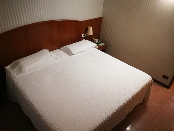 Un lit bien fait, avec du linge de lit blanc et des oreillers, une tête de lit en bois, une lampe de chevet et une table de chevet avec un téléphone.