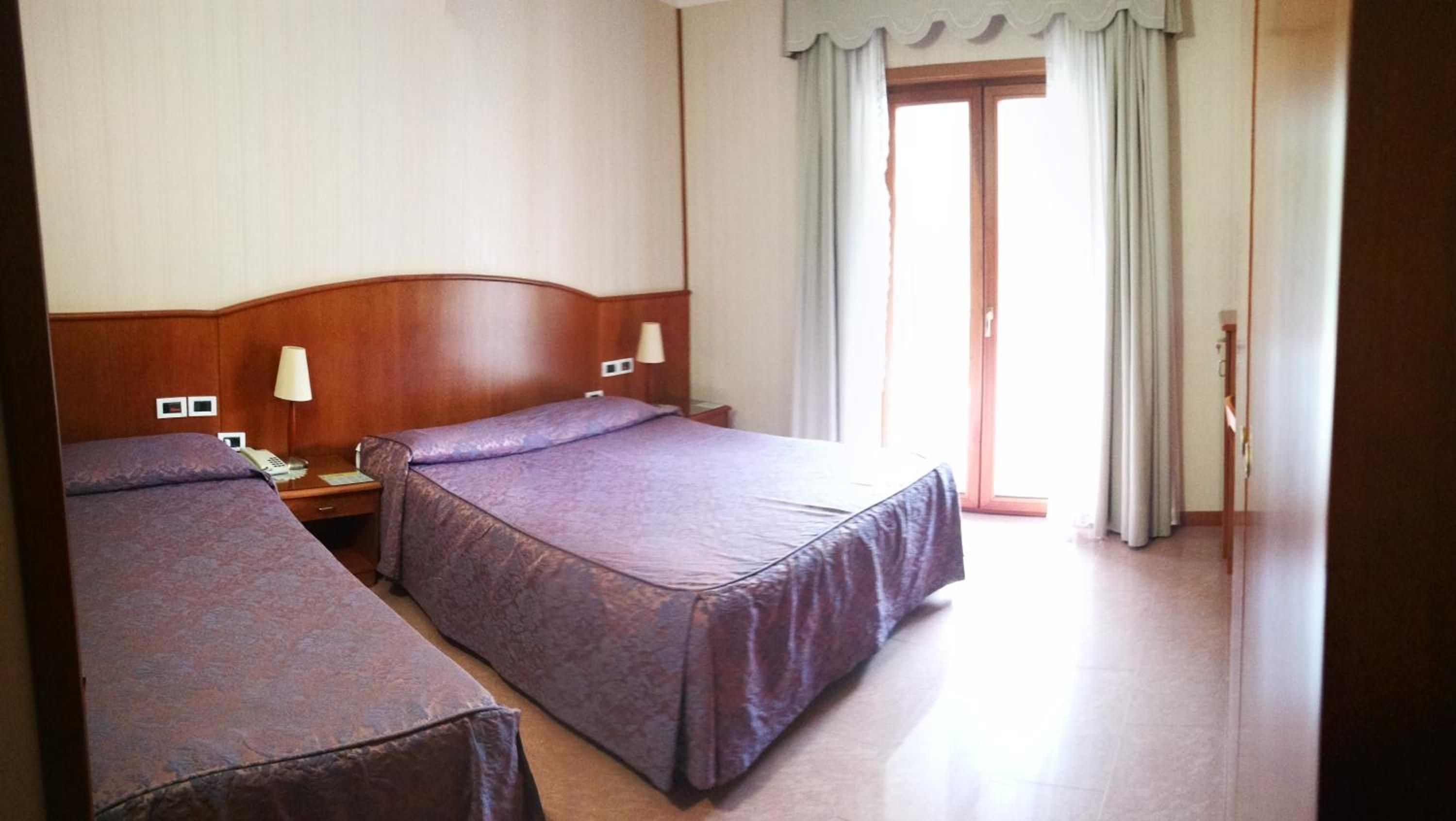 Ein Hotelzimmer mit zwei Betten, einem Holzkopfteil, Nachttischlampen, einem Telefon und einer Tür zu einem Balkon.