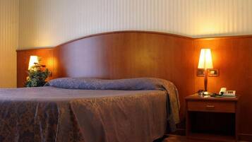 Ein Hotelzimmer mit einem gebogenen Kopfteil, einem Bett mit gemustertem Bettbezug, einem Nachttisch mit Lampe und einem kleinen Tisch mit Telefon.