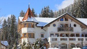 A large, multi-story building with a snow-covered roof, surrounded by snow-covered trees and a stone wall. - Turmhotel Gschwendt (Valle di Casies)