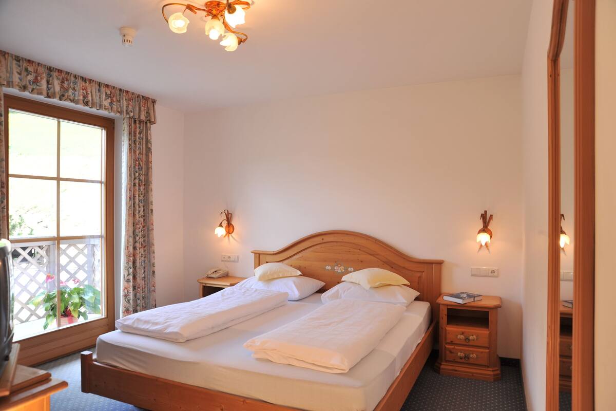 A double bed with two pillows, a wooden headboard, bedside tables with lamps, a window with curtains, and a small plant.