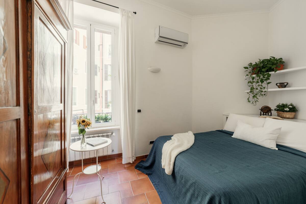 Un dormitorio con cama, ventana con cortinas, radiador, una mesita con un florero y un aire acondicionado instalado en la pared.