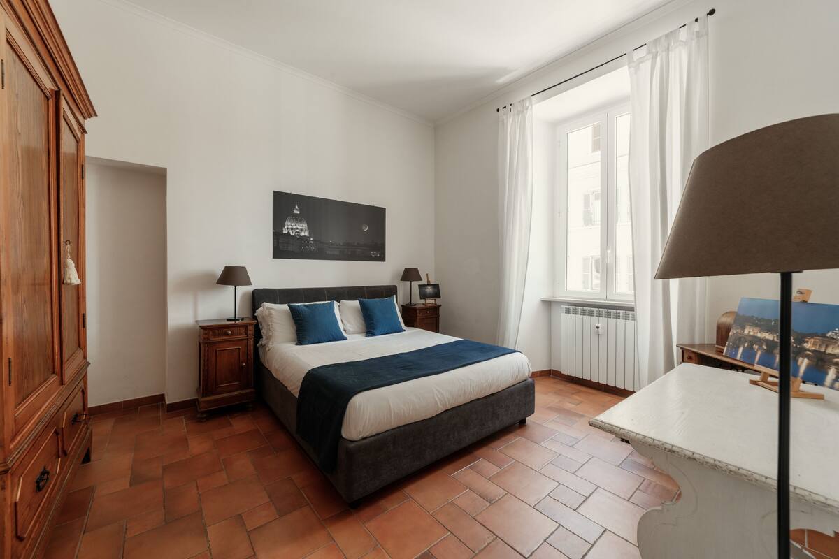 Un dormitorio con una cama, mesitas de noche, un cuadro, una ventana con cortinas y un radiador.