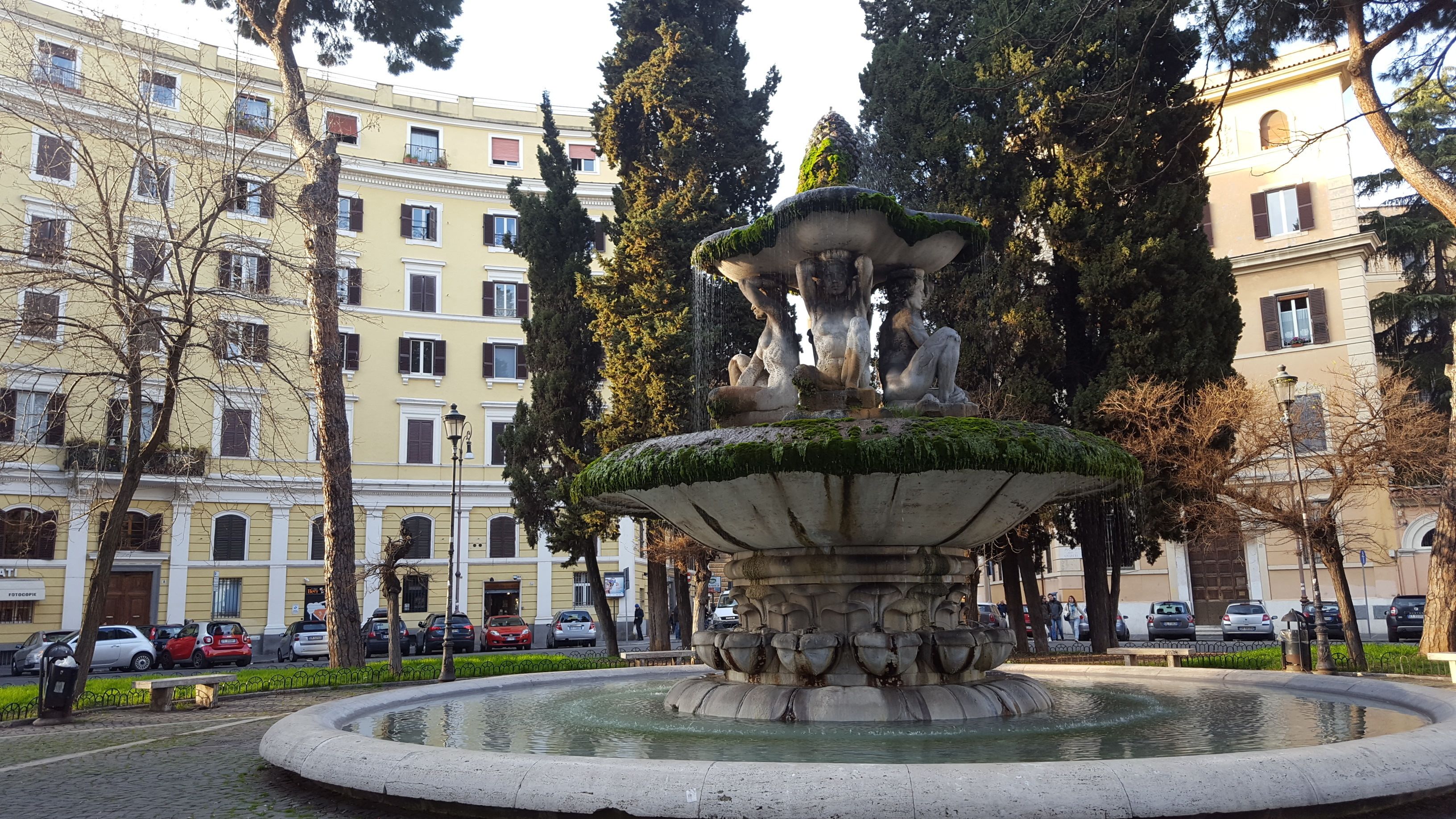 Une fontaine ornée de statues, située sur une place publique et entourée d’immeubles.