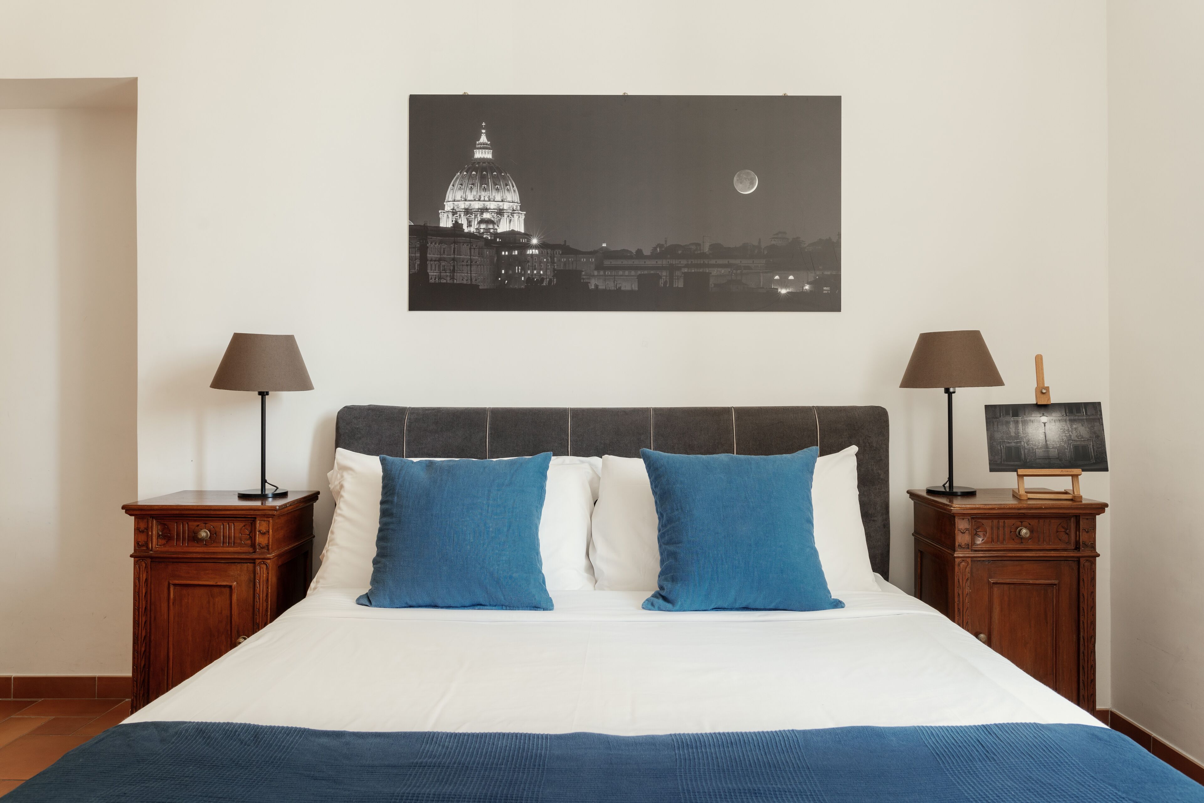 Un lit bien fait, avec des oreillers bleus et blancs, deux tables de chevet avec des lampes, et un tableau représentant un paysage urbain encadré au mur.