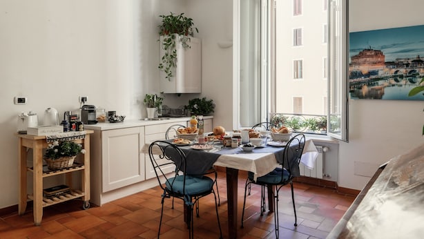 A dining area with a table set for two, a white cabinet with a hanging plant, and a window with a view of a building. - Domus Quiritum (Rome)