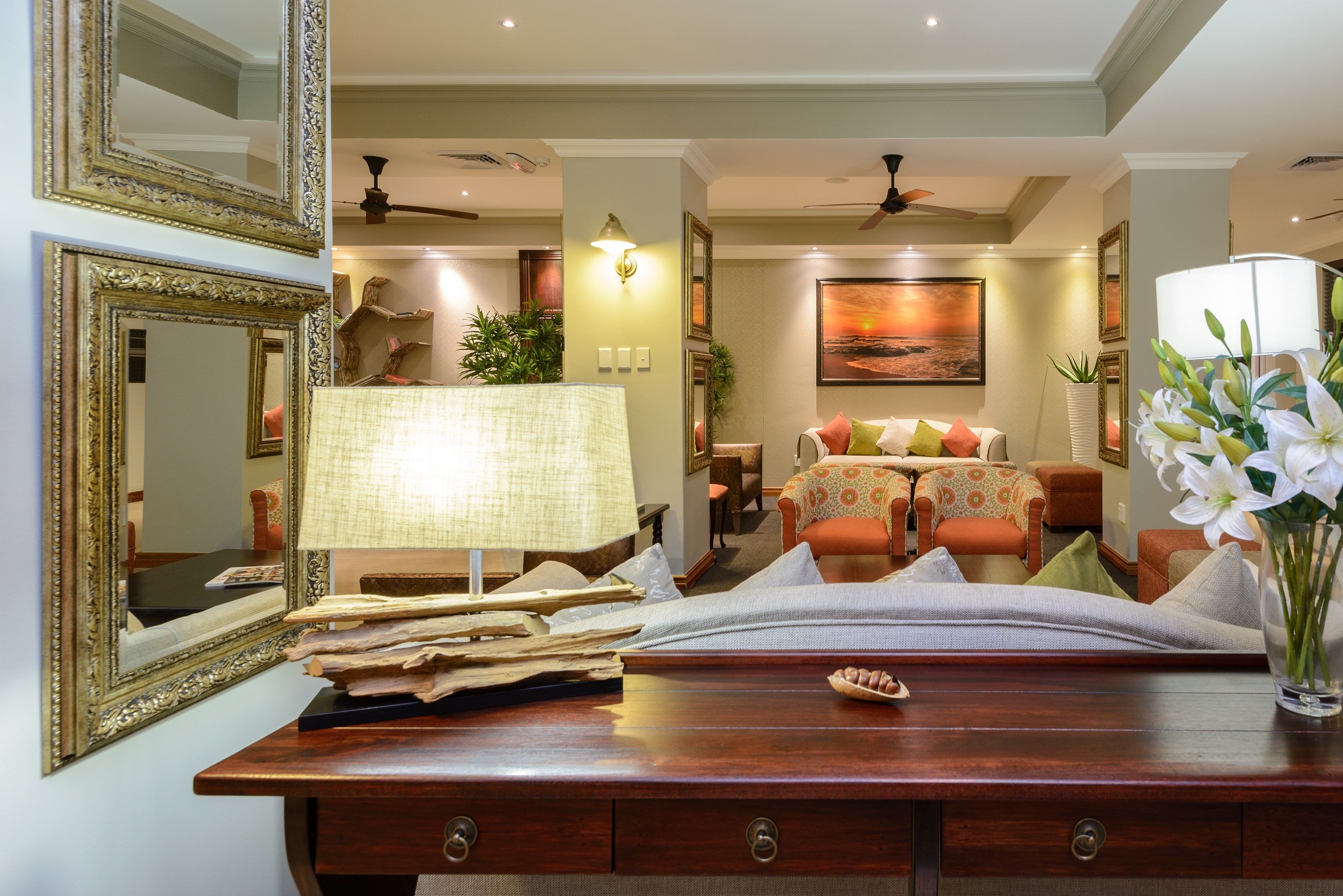 A well-lit living room with a wooden coffee table, a lamp, and a sofa with cushions. There are framed pictures on the wall and a large painting above the sofa.