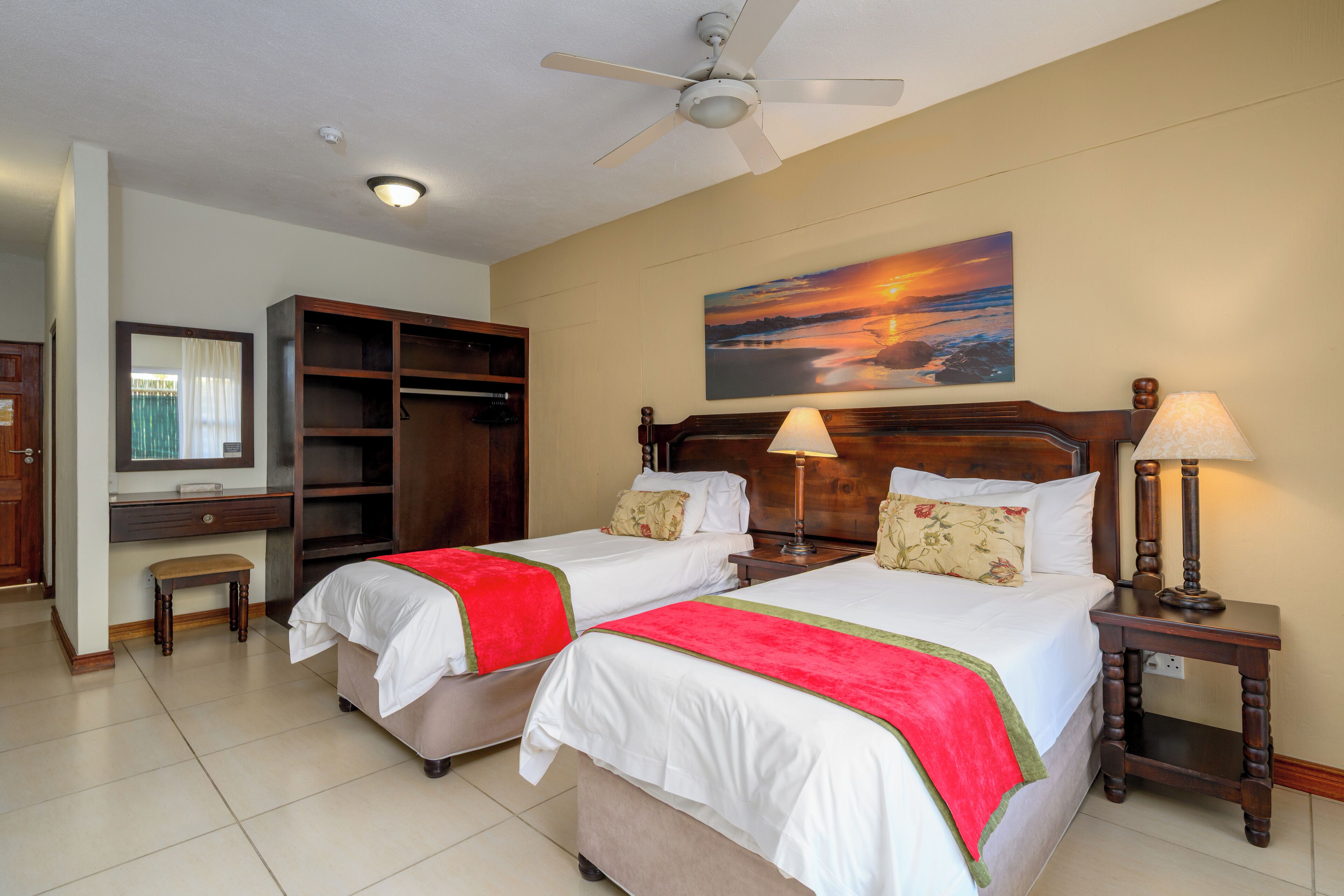 A hotel room with two beds, a desk, a wardrobe, and a painting of a sunset.