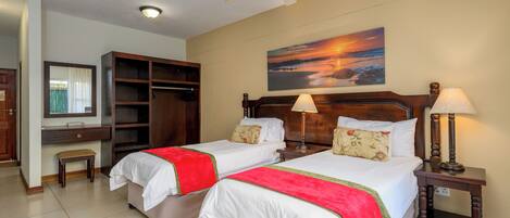 A hotel room with two beds, a desk, a wardrobe, and a painting of a sunset.
