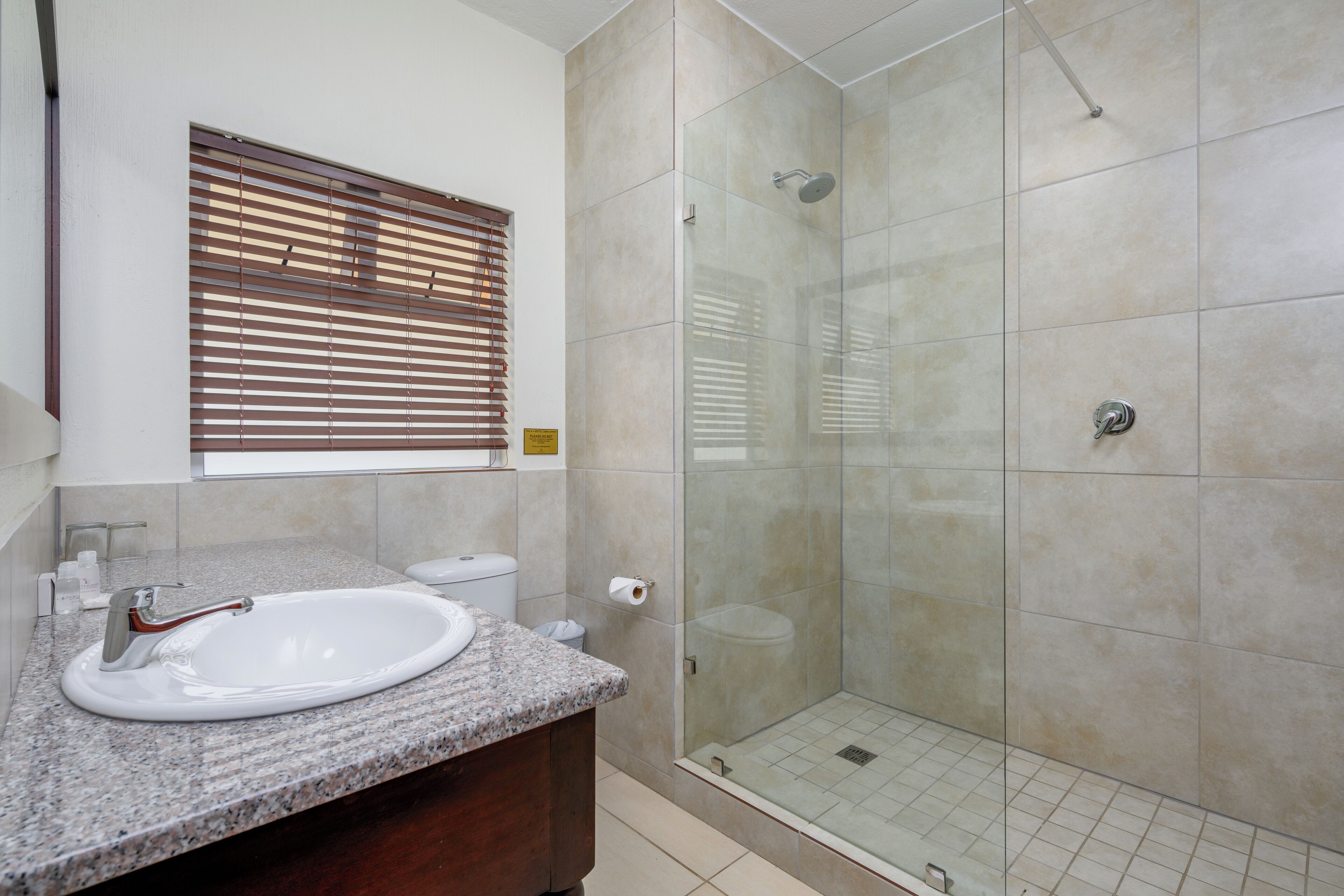 A bathroom with a granite countertop, a white sink, a glass-enclosed shower, and a window with blinds.