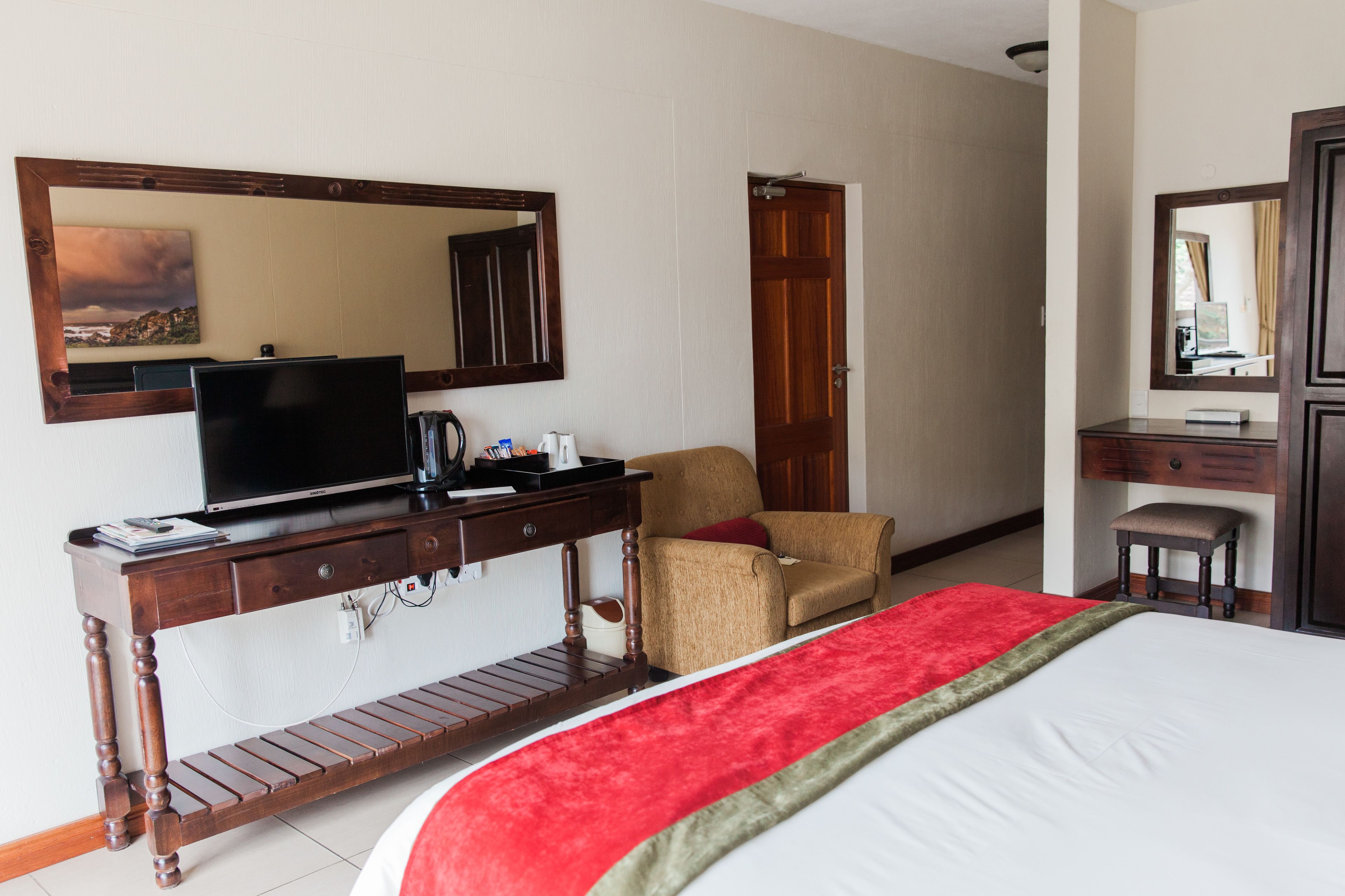 A hotel room with a bed, a television on a wooden console, a chair, a desk, and a mirror.