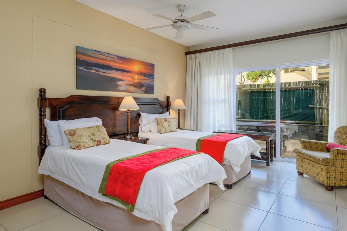 A hotel room with two beds, a ceiling fan, a painting of a sunset, a sliding glass door to a balcony, and a wicker chair.