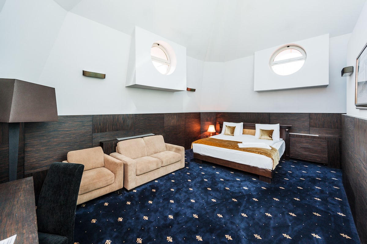 A hotel room with a large bed, a sofa, a desk, and two windows.