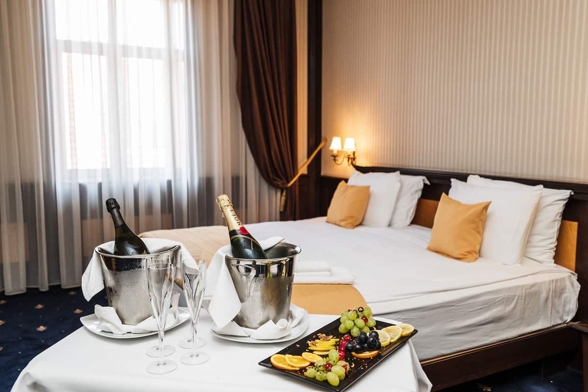 A hotel room with a bed, a nightstand with a bottle of champagne, two glasses, and a fruit platter.