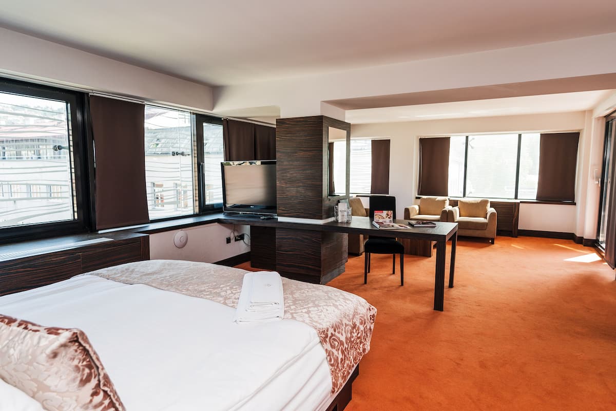 A hotel room with a large bed, a desk, a chair, and a sofa. There is a window with curtains and a view of the outside.