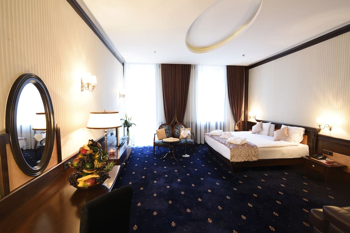 A hotel room with a large bed, a round table with chairs, a small round table with a lamp, a mirror, and a fruit bowl.
